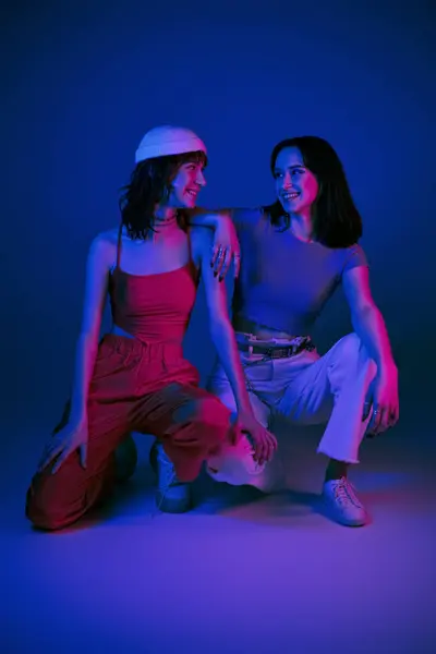 Счастливые молодые девушки позируют в стильной одежде и смотрят друг на друга под фиолетовыми огнями — стоковое фото