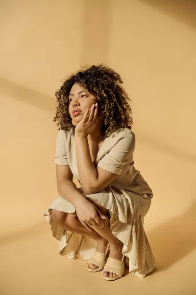 Una hermosa joven afroamericana con el pelo rizado sentada en el suelo con la mano en la cara, perdida en un profundo pensamiento. - foto de stock