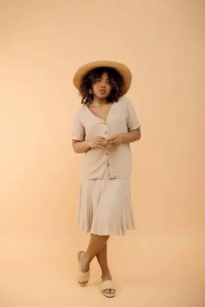 Una hermosa joven afroamericana con el pelo rizado posando elegantemente en un vestido de verano floral y un elegante sombrero de ala ancha. - foto de stock