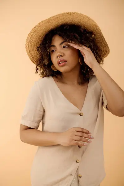 Una elegante mujer afroamericana con el pelo rizado da una pose en un sombrero de paja para un retrato en un ambiente sereno estudio. - foto de stock