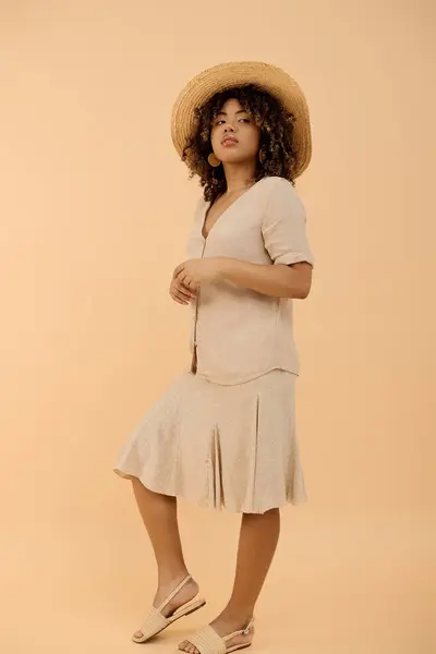 A fashionable young African American woman with curly hair posing confidently in a summer dress and hat in a studio setting. — Stock Photo