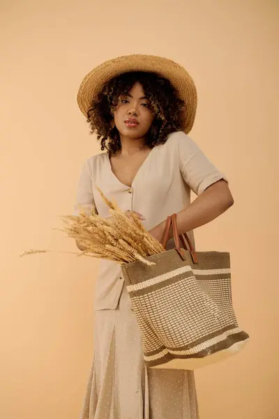 Joven mujer afroamericana con el pelo rizado en un sombrero de paja con elegancia sosteniendo una bolsa en un entorno de estudio brillante. - foto de stock