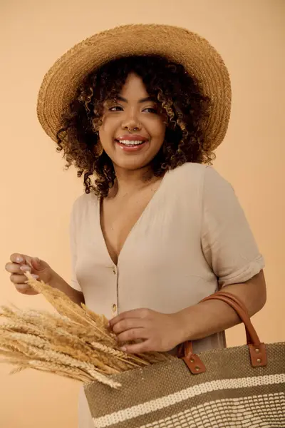 Eine elegante Afroamerikanerin mit lockigem Haar, gekleidet in ein Sommerkleid, eine Tasche in der Hand, während sie einen Strohhut trägt. — Stockfoto