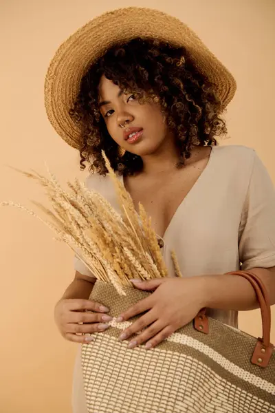 Belle femme afro-américaine dans un chapeau de paille tenant un sac élégant, exsudant élégance et sophistication dans un cadre de studio. — Photo de stock