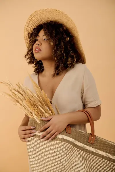 Joven mujer afroamericana con el pelo rizado, con un sombrero de paja, sosteniendo una bolsa en un elegante ambiente de estudio. - foto de stock