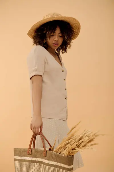 Une jeune femme afro-américaine en robe d'été tenant un chapeau de paille et un sac, émanant élégance et style. — Photo de stock