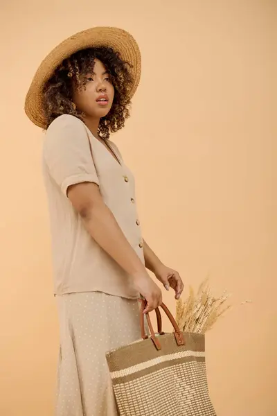 Une belle jeune afro-américaine aux cheveux bouclés tenant un sac tout en portant un chapeau de paille dans un décor de studio. — Photo de stock