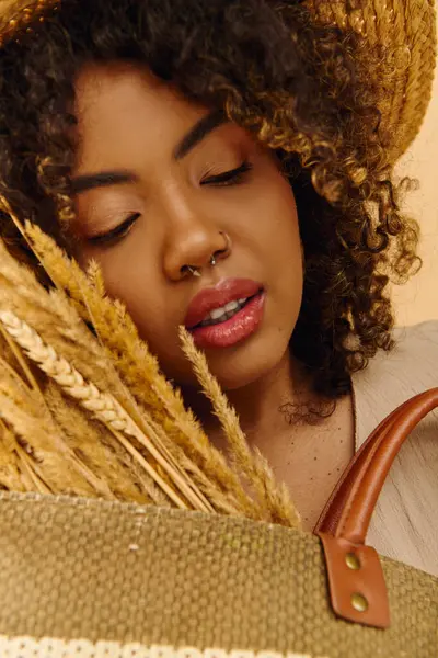Una hermosa joven afroamericana con el pelo rizado sostiene elegantemente una bolsa marrón mientras usa un sombrero de paja en un entorno de estudio. - foto de stock