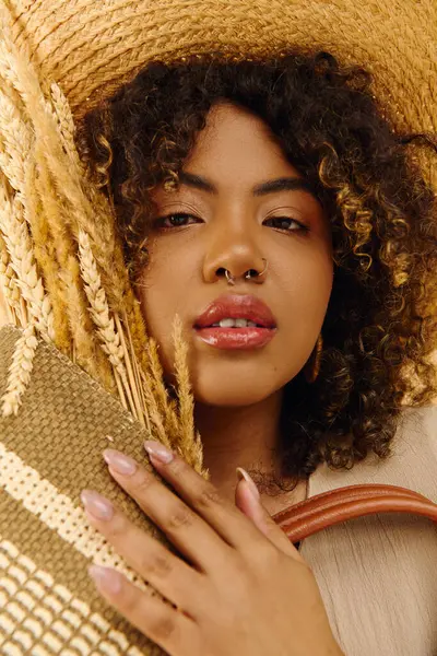 Una hermosa joven afroamericana con el pelo rizado usando un sombrero de paja y sosteniendo un bolso en un ambiente de estudio. - foto de stock