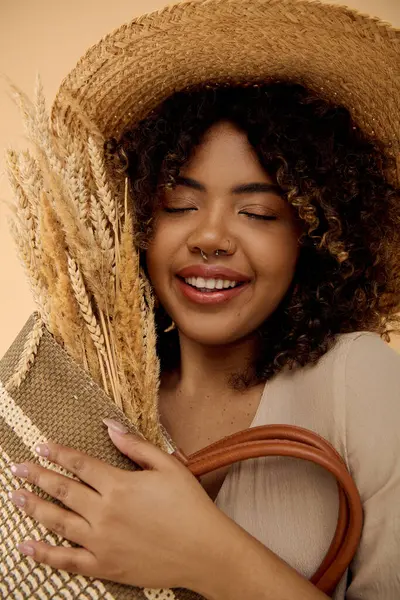 Hermosa mujer afroamericana con el pelo rizado, usando un sombrero de paja, y sosteniendo un bolso en un ambiente de estudio. - foto de stock