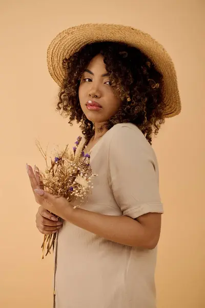 Une belle jeune femme afro-américaine aux cheveux bouclés portant un chapeau de paille, tenant un bouquet de fleurs dans un moment serein. — Photo de stock