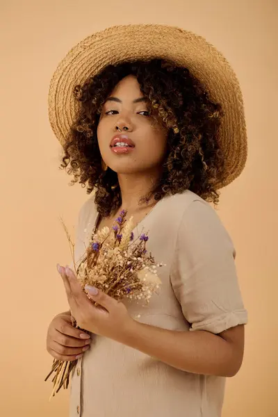 A beautiful young African American woman with curly hair, wearing a straw hat and holding flowers in a studio setting. — Stock Photo