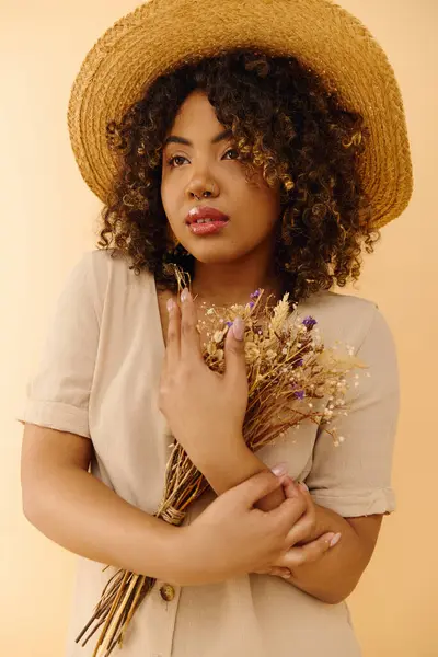 Una hermosa mujer afroamericana con el pelo rizado, en un vestido de verano, sosteniendo flores y usando un sombrero de paja. - foto de stock