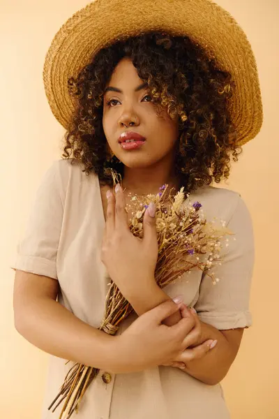 A beautiful African American woman with curly hair, in a summer dress, holding flowers and wearing a straw hat. — Stock Photo