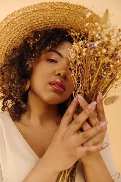 Une jeune femme afro-américaine aux cheveux bouclés dans une robe d'été, tenant un bouquet de fleurs colorées tout en portant un chapeau de paille élégant. — Photo de stock