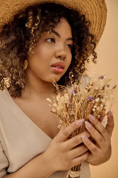 Una hermosa joven afroamericana con el pelo rizado, vestida con un vestido de verano, sosteniendo un ramo de flores secas. - foto de stock