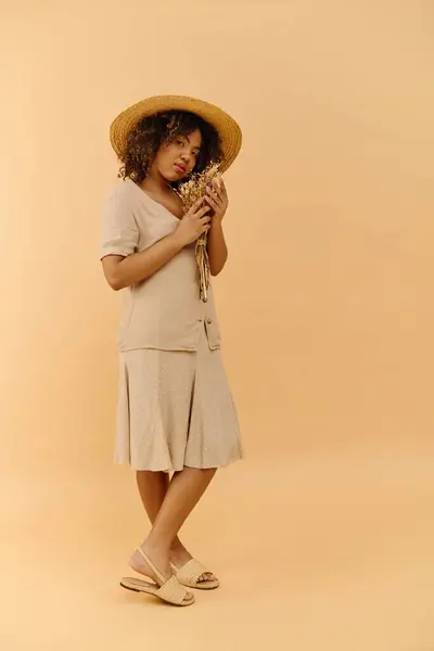 Una hermosa joven afroamericana con el pelo rizado sostiene una flor mientras usa un sombrero elegante en un ambiente de estudio. - foto de stock