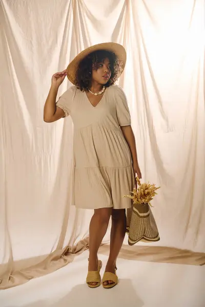 A beautiful young African American woman with curly hair poses gracefully in a stylish dress and hat in a studio setting. — Stock Photo