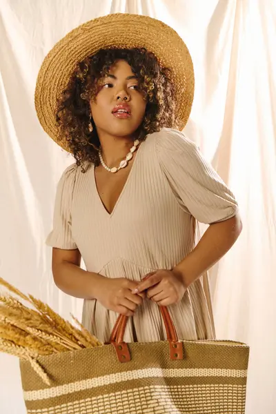 A young African American woman with curly hair is elegantly posing in a straw hat while holding a straw bag in a studio setting. — Stock Photo
