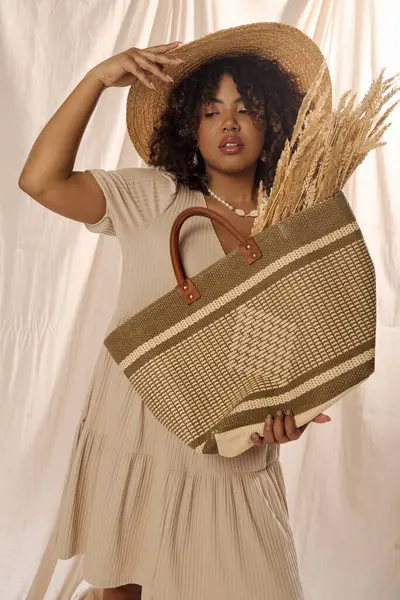 Beautiful African American woman with curly hair in a white dress, gracefully holding a stylish straw bag. — Stock Photo