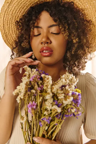 Une belle jeune femme afro-américaine aux cheveux bouclés portant un chapeau de paille, tenant un bouquet de fleurs vibrantes dans un cadre de studio. — Photo de stock