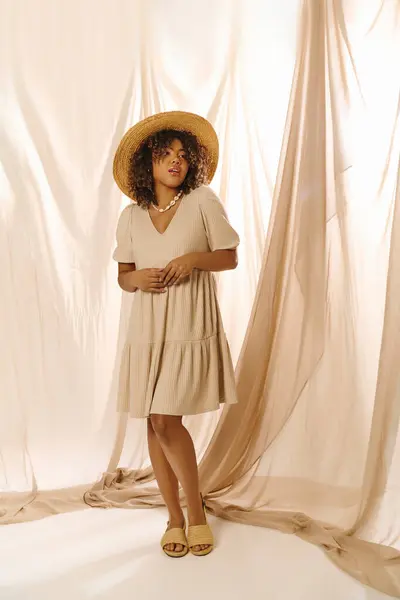 Une jeune afro-américaine élégante aux cheveux bouclés, coiffée d'un chapeau, se dresse en toute confiance sur fond blanc dans un studio. — Photo de stock