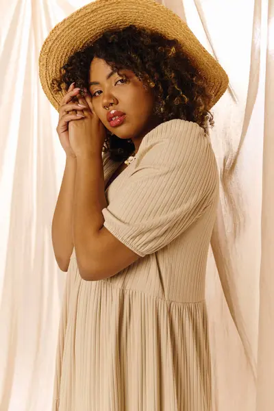 Une jeune femme afro-américaine glamour aux cheveux bouclés portant une robe d'été et un chapeau pose élégamment pour un portrait. — Photo de stock