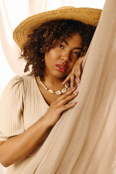 A beautiful young African American woman with curly hair in a straw hat is peeking out from behind a curtain in a studio setting. — Stock Photo