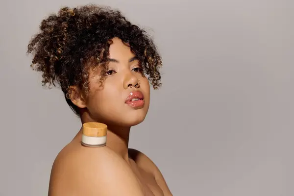 A beautiful young African American woman with curly hair with cream jar on shoulder, focusing on enhancing her skins beauty. — Stock Photo