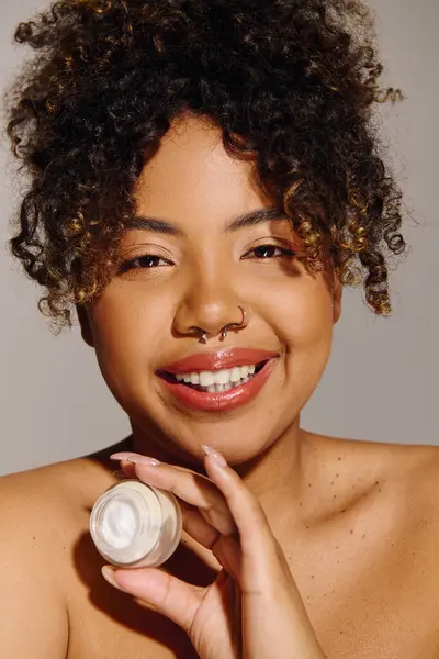 Beautiful African American woman with curly hair holding a jar of cream in a studio setting, focusing on skincare. — Stock Photo