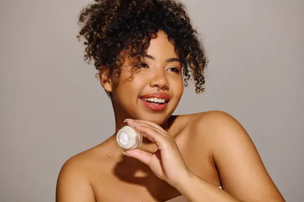 A beautiful young African American woman with curly hair holding a jar of cream in a studio setting, focused on skin care and beauty. — Stock Photo