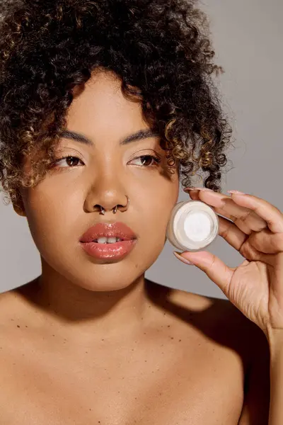 A young African American woman with curly hair holds a jar of cream in front of her face, emphasizing skincare and beauty. — Stock Photo