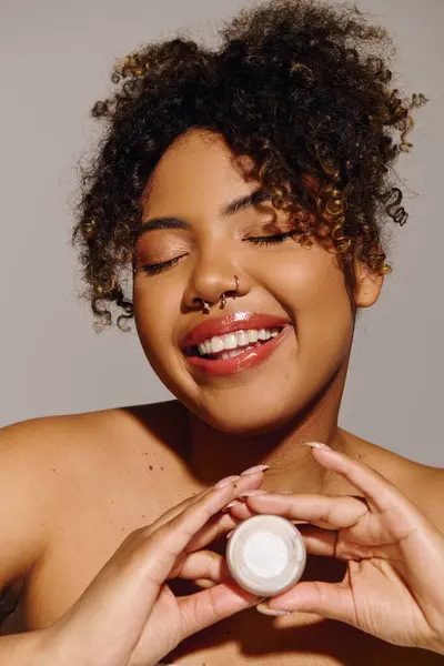 Красивая афроамериканка держит банку сливок с вьющимися волосами, подчеркивая уход за кожей и красоту рутины. — стоковое фото