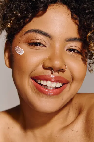 Uma bela jovem afro-americana com cabelo encaracolado mostrando um creme branco na bochecha em um estúdio. — Fotografia de Stock