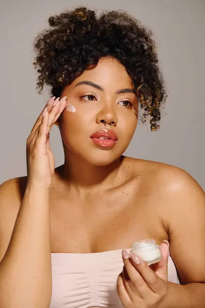Eine junge Afroamerikanerin mit lockigem Haar hält sich ein Glas Creme vor das Gesicht. — Stockfoto