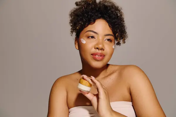 A stunning African American woman with curly hair, wrapped in a white towel, applying cream from a jar. — Stock Photo