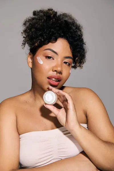 Uma bela jovem afro-americana com cabelo encaracolado segurando um frasco de creme, irradiando confiança e beleza. — Fotografia de Stock