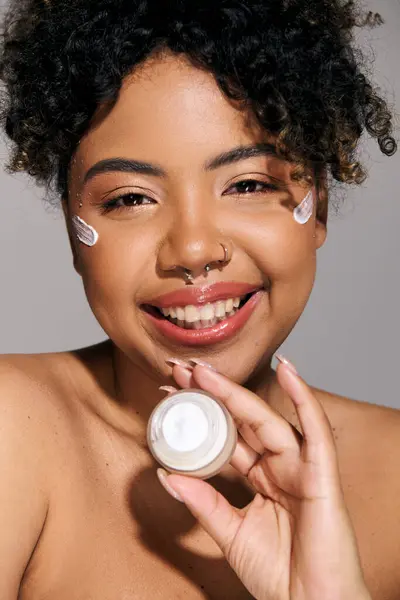 Joven mujer afroamericana con el pelo rizado aplicando crema de un frasco a su cara en un ambiente de estudio. - foto de stock