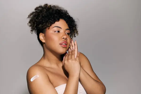 A young African American woman with curly hair in a strapless top delicately holds her hand to her face in a studio setting. — Stock Photo