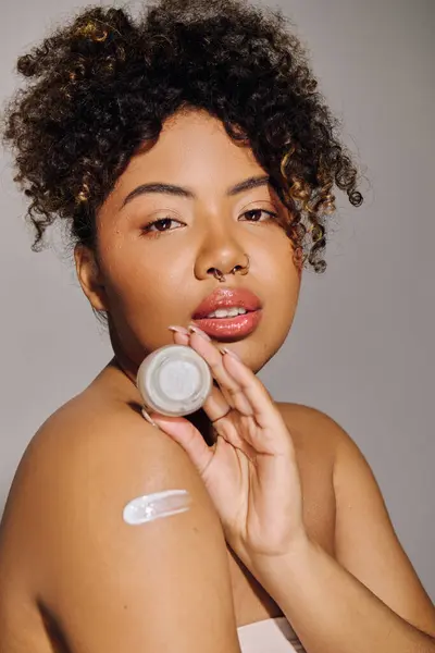 Young African American woman with curly hair in a studio setting, applying a generous amount of cream onto her face. — Stock Photo