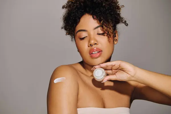 Jeune femme afro-américaine aux cheveux bouclés applique délicieusement de la crème sur son visage dans un cadre de studio. — Photo de stock