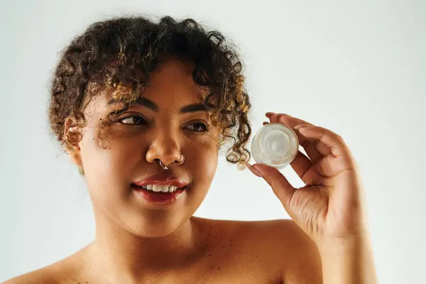 Una donna afroamericana tiene elegantemente una bottiglia di crema nella mano destra su uno sfondo vibrante. — Foto stock