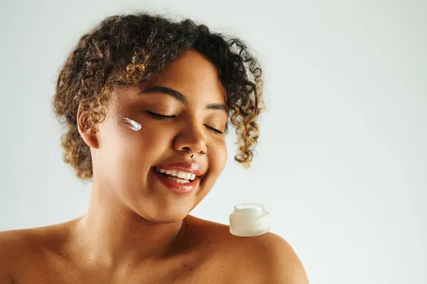 Cheerful African American woman smiling while holding a cream bottle. — Stock Photo