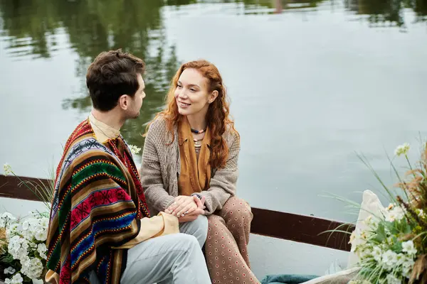 A man and a woman, dressed in boho style clothes, peacefully sit on a boat in a lush green park setting. — Stock Photo