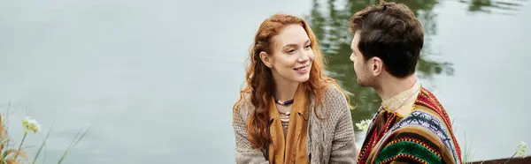 Un hombre y una mujer vestidos con ropa de estilo boho se sientan en un banco junto a un tranquilo cuerpo de agua, disfrutando de una cita romántica en un parque verde. — Stock Photo