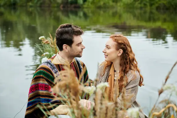 A man and woman in boho attire relax by a serene body of water, enjoying a romantic date in a lush green park setting. — Stock Photo