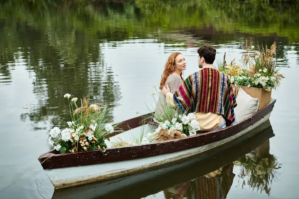 Un hombre y una mujer en ropa de estilo boho reman en un barco lleno de flores vibrantes en un exuberante parque verde, disfrutando de una cita romántica. - foto de stock