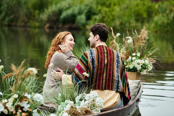 A couple in boho clothing navigate a boat filled with vibrant flowers in a serene green park setting. — Stock Photo