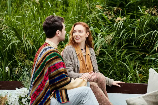A man and a woman in boho style clothes are seated on a bench, engaged in conversation in a lush green park setting. — Stock Photo