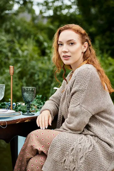 A woman in boho style apparel gracefully sits next to a wine glass at a table in a lush green park. — Stock Photo